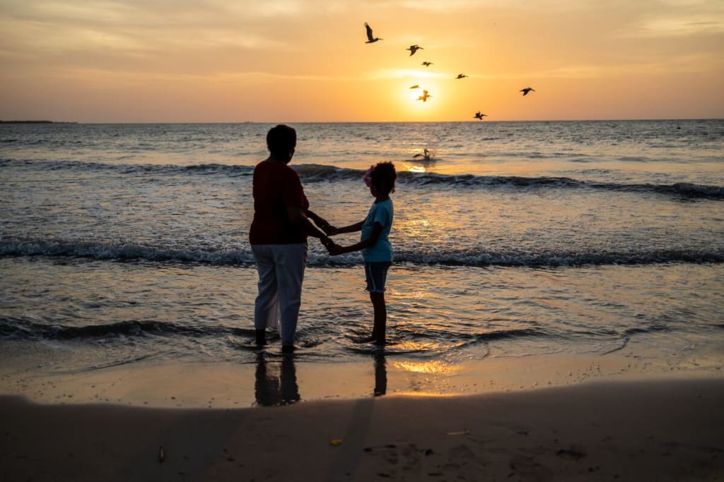 A woman and a girl hold hands in front of a sunset over water.